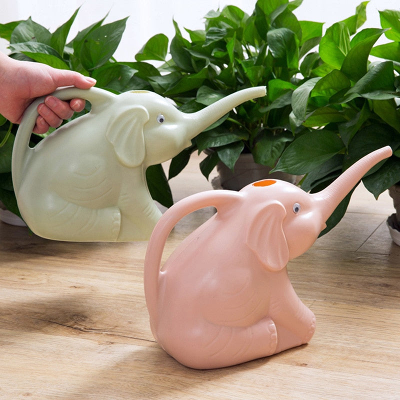 Two elephant-shaped watering cans, one green and one pink, placed on a wooden floor with a background of lush green potted plants. A hand holds the green can, demonstrating its handle and spout design.